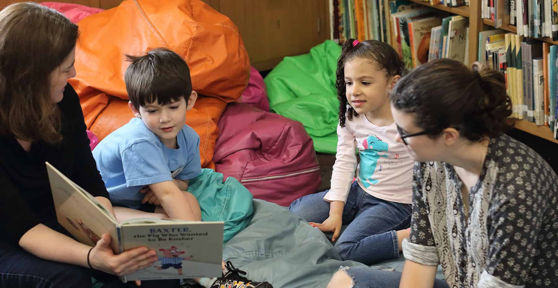 An image of a woman with short brown hair interacting and reading a story to 3 children who sit around, smiling and listening to the book.