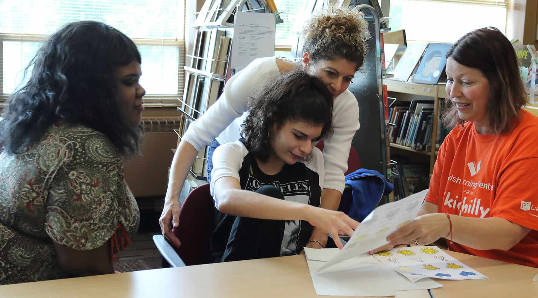 Three adults sit around a girl and help her with a worksheet. They are all smiling and engaged in the work.